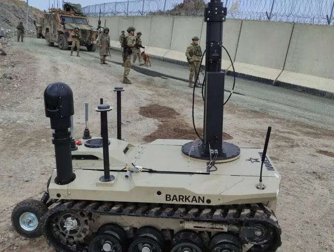 Tan BARKAN UGV fitted with two raised mast gimbals, soldiers and vehicles behind a wall.