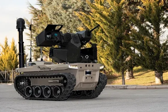 Side view of a tan BARKAN tracked UGV with raised gimbal mast parked on paved ground.
