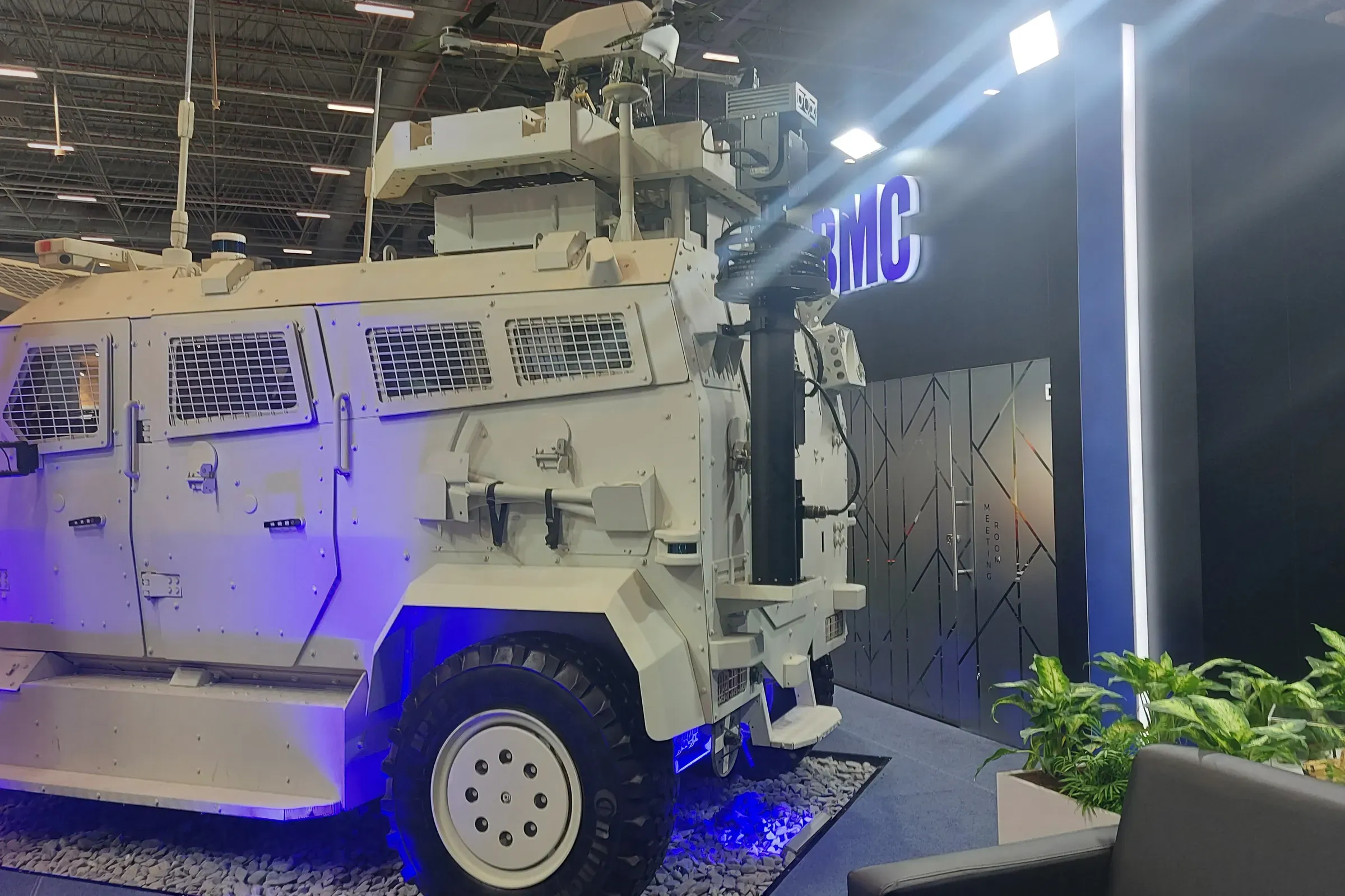 White armoured 4x4 vehicle displayed on a stand with a mast raised behind the cab, blue floor lighting.