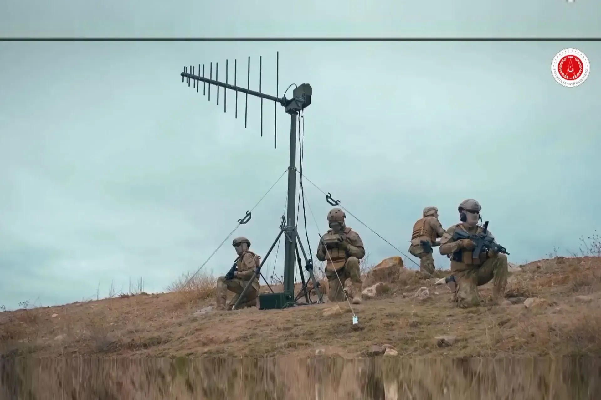 Soldiers kneeling on a hilltop beside a guyed mast carrying a yagi antenna array under overcast sky.