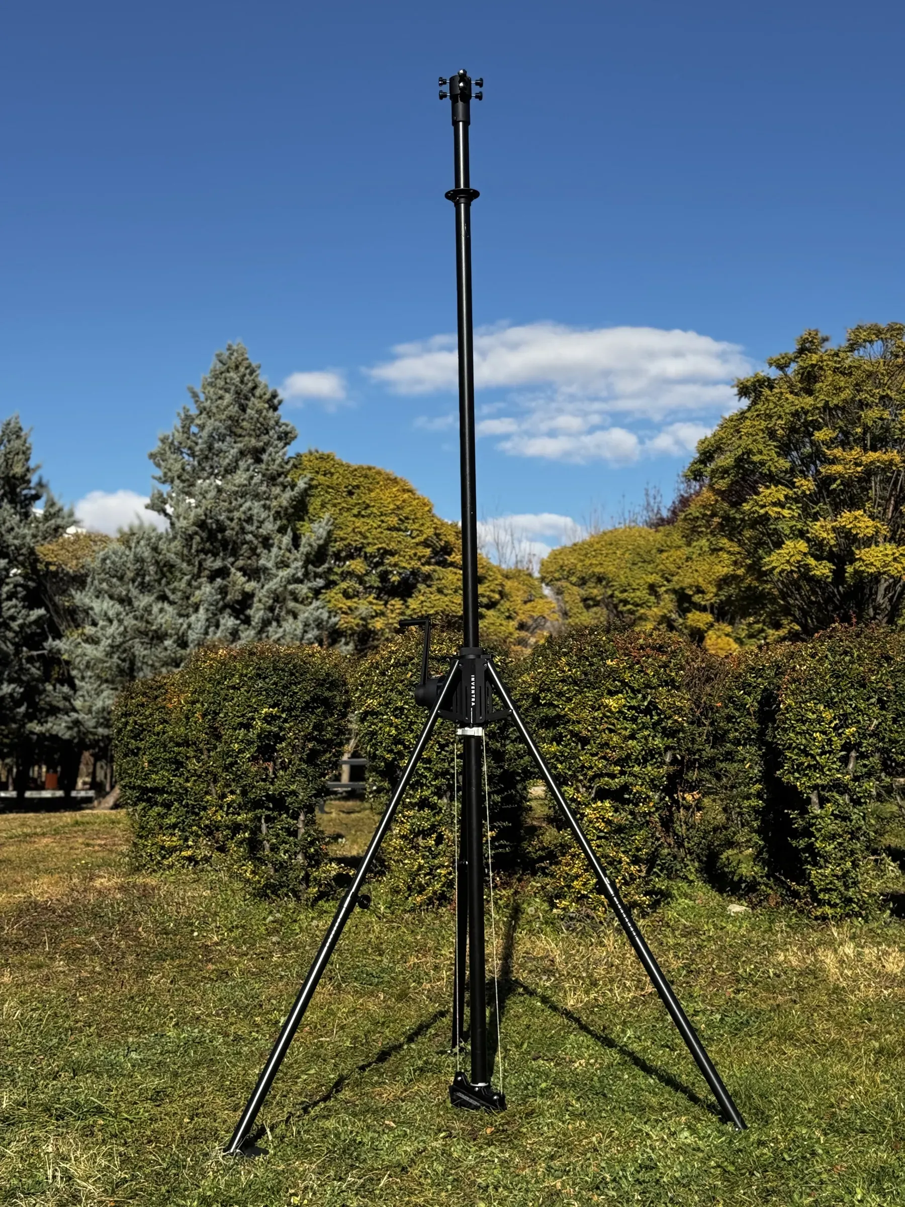 Portable tripod mast fully extended on grass against blue sky and autumn trees.