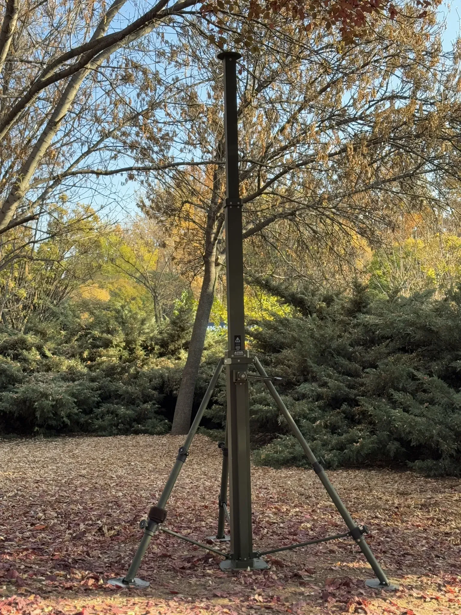 Manual mast fully extended on tripod in autumn woodland, viewed from ground among bare trees.