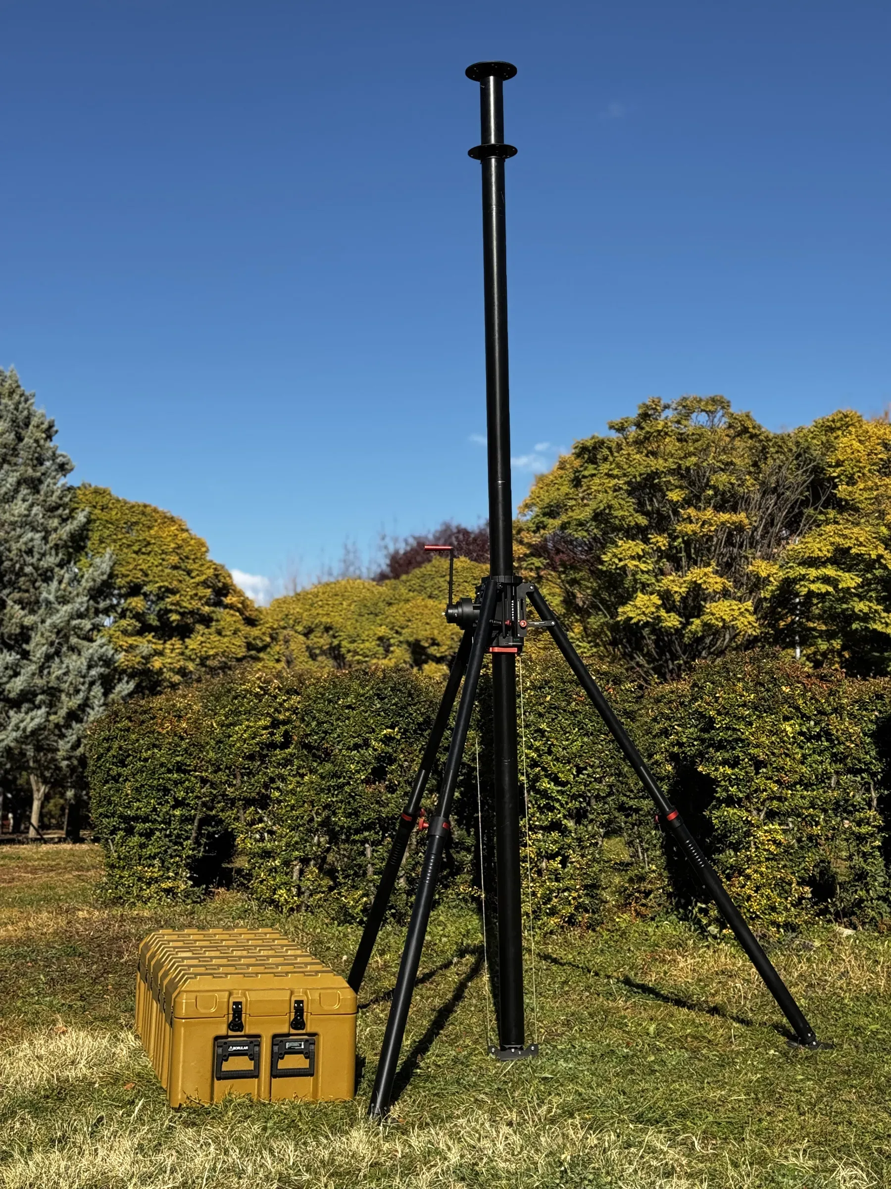 Extended tripod mast standing on grass in front of shrubs, olive transit case at its foot.