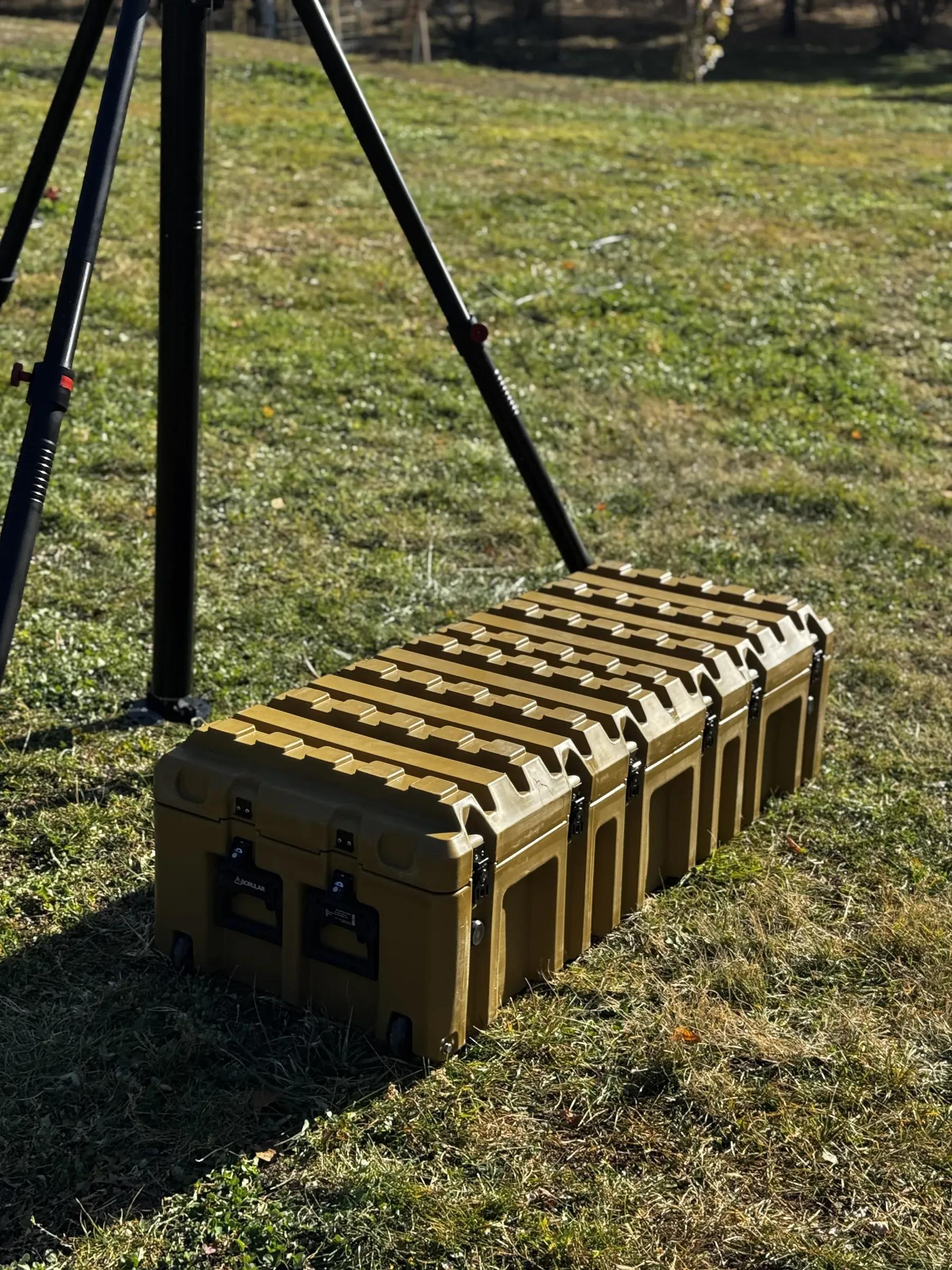 Tripod legs planted on grass beside a closed olive-green ruggedised transit case for the system.
