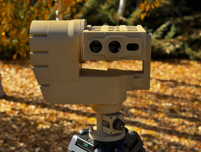 Tan PED12 positioner with multi-aperture sensor head on a black tripod, standing on autumn leaf litter among birch trees.