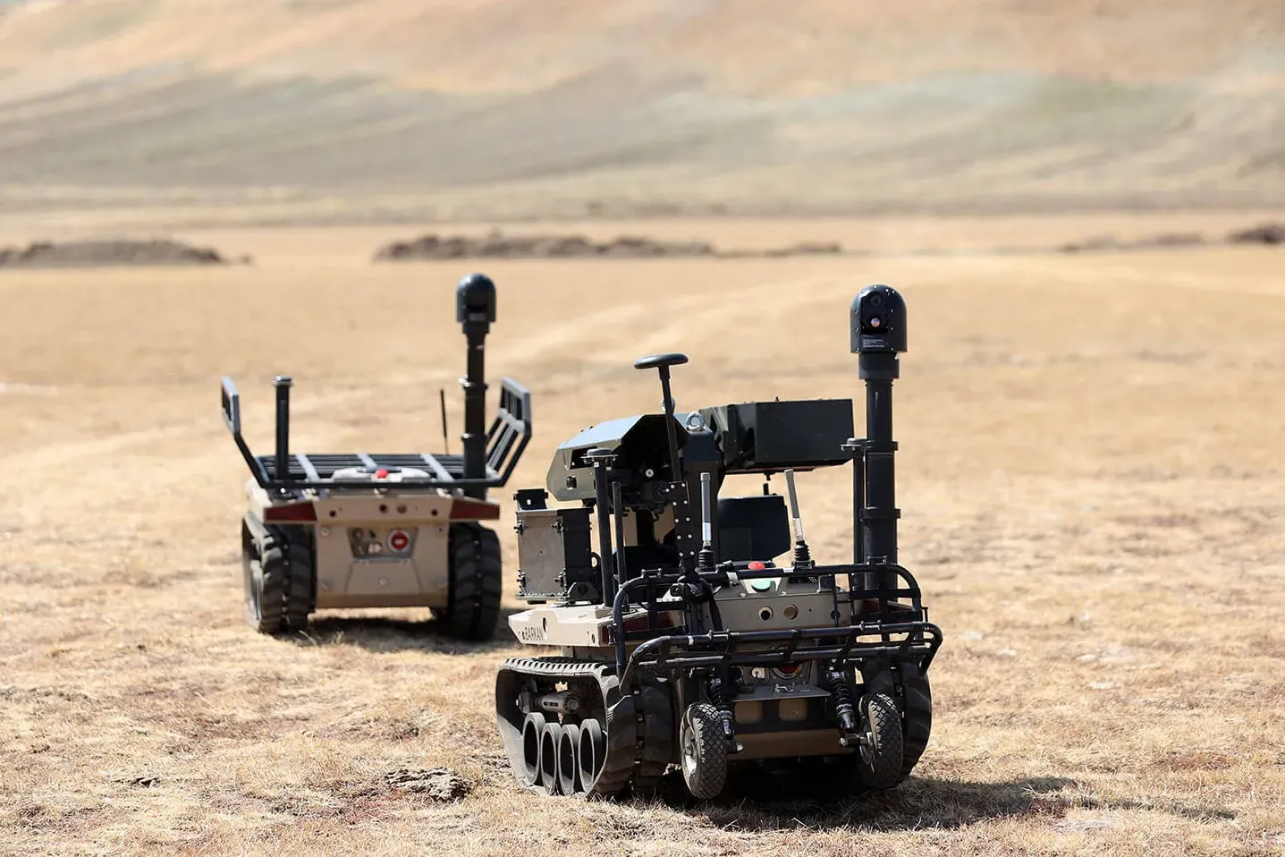 Two unmanned ground vehicles parked on open steppe, the nearer one carrying an extended mast and sensor head.