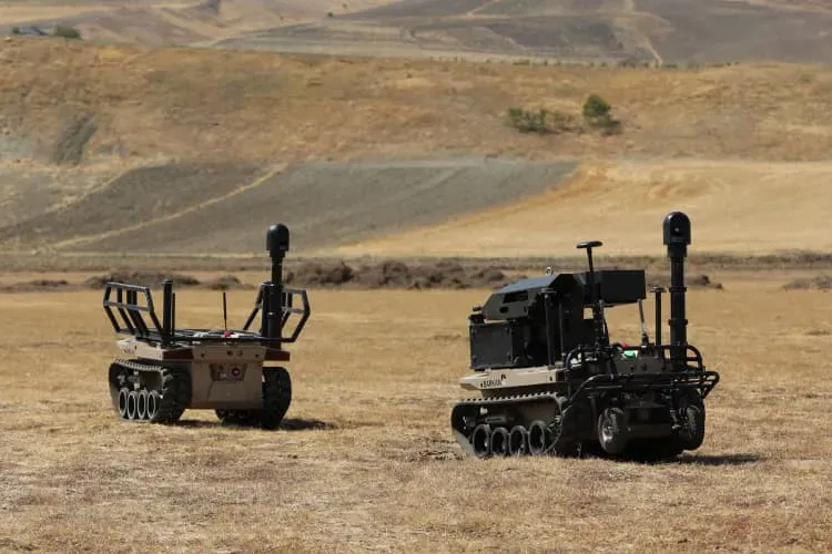 Two unmanned ground vehicles on a dry field, the right-hand one carrying an extended mast and sensor head.