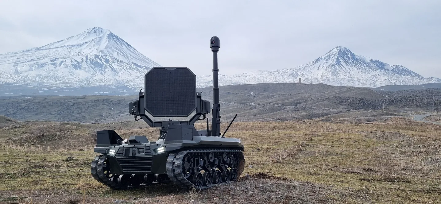 A tracked unmanned ground vehicle on open ground before snow-covered mountains, carrying a large acoustic panel with a telescopic mast and a stabilised gimbal raised behind it.