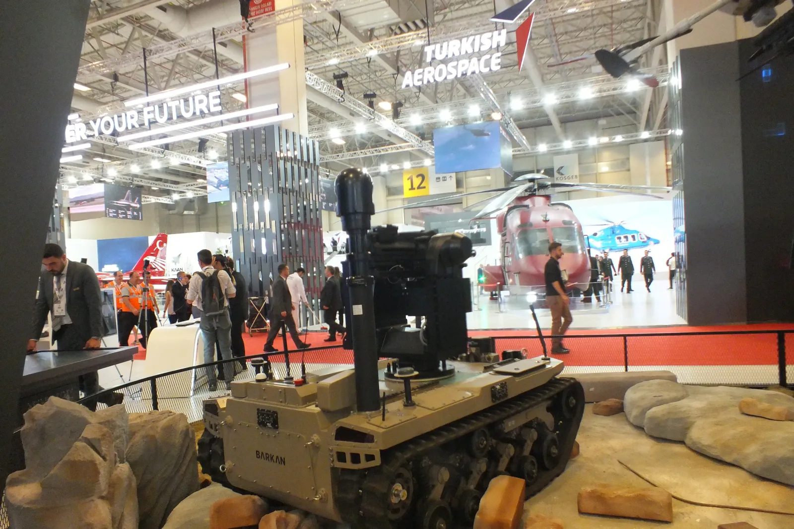 A tracked unmanned ground vehicle on the stand with a telescopic mast raised and a stabilised gimbal at its top.