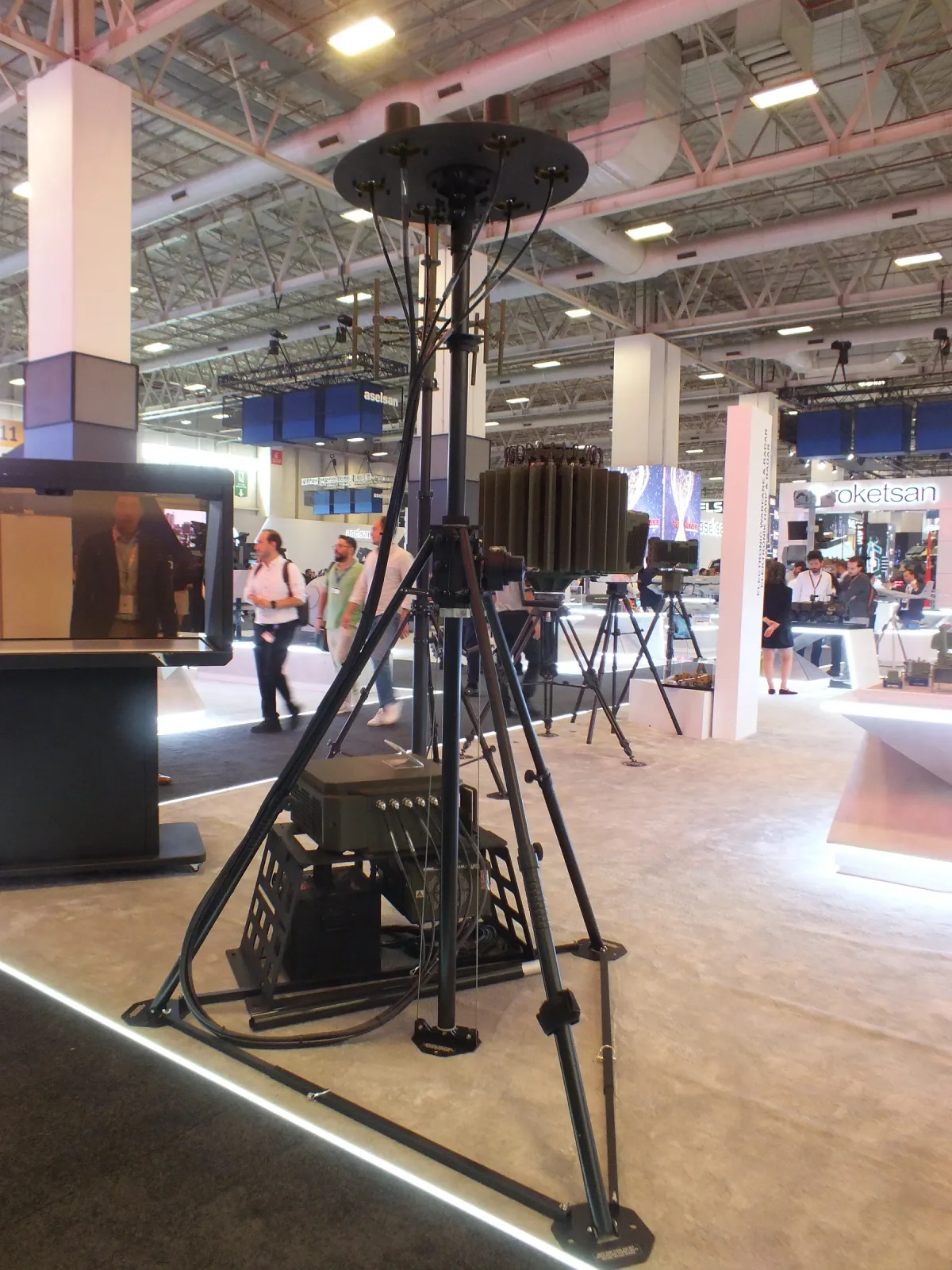 A portable tripod mast set up on the exhibition floor with its guy rings and payload head.