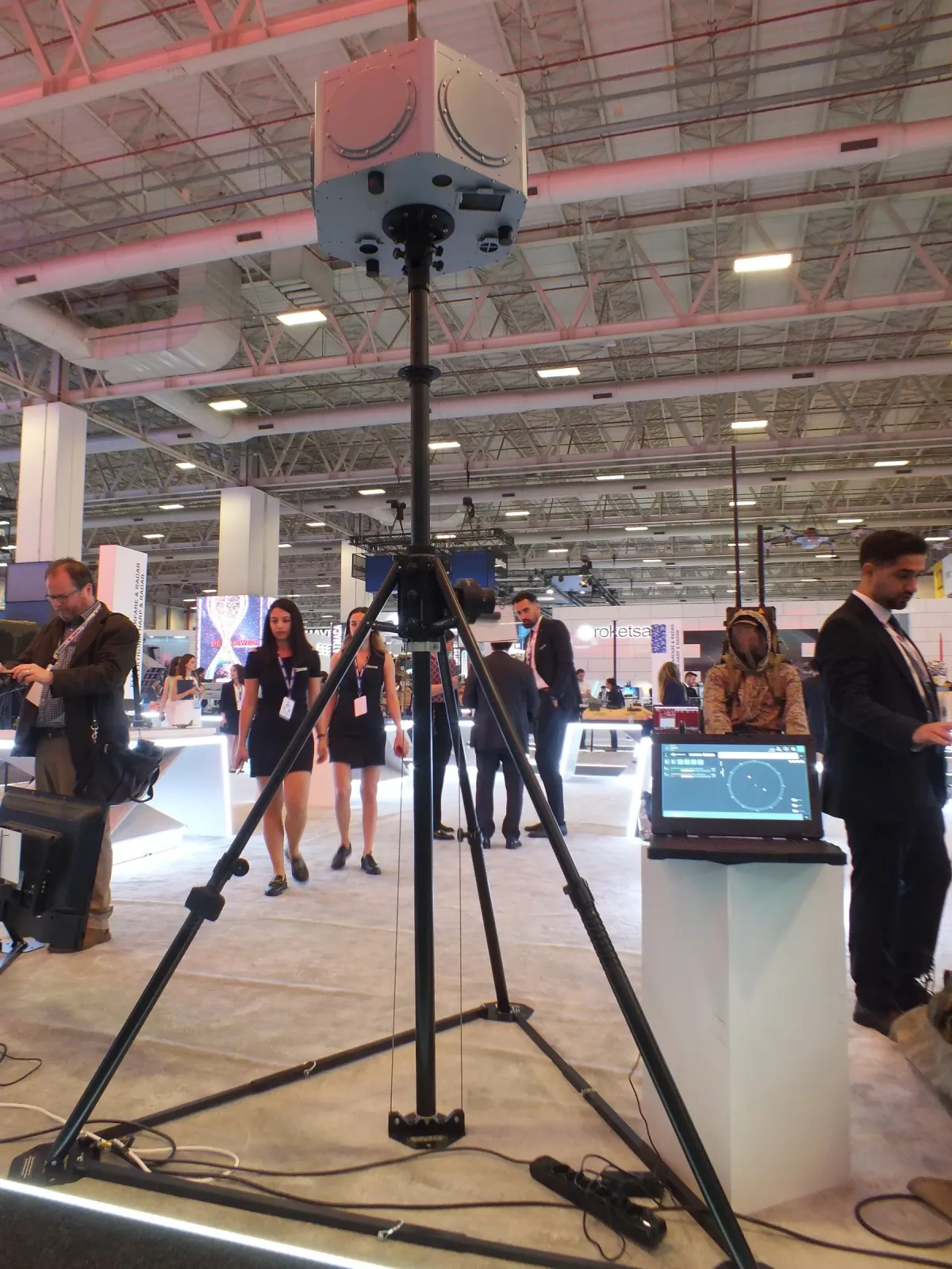 A portable tripod mast set up on the exhibition floor with its guy rings and payload head.