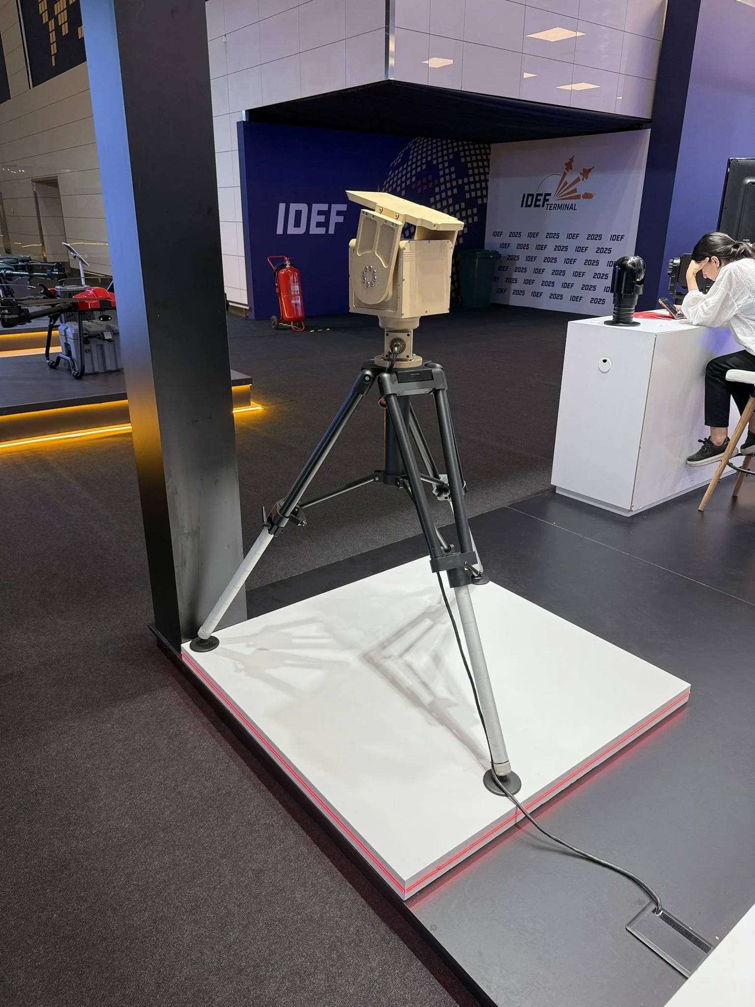Sand-coloured pan-tilt unit on a black tripod, standing on a white plinth.