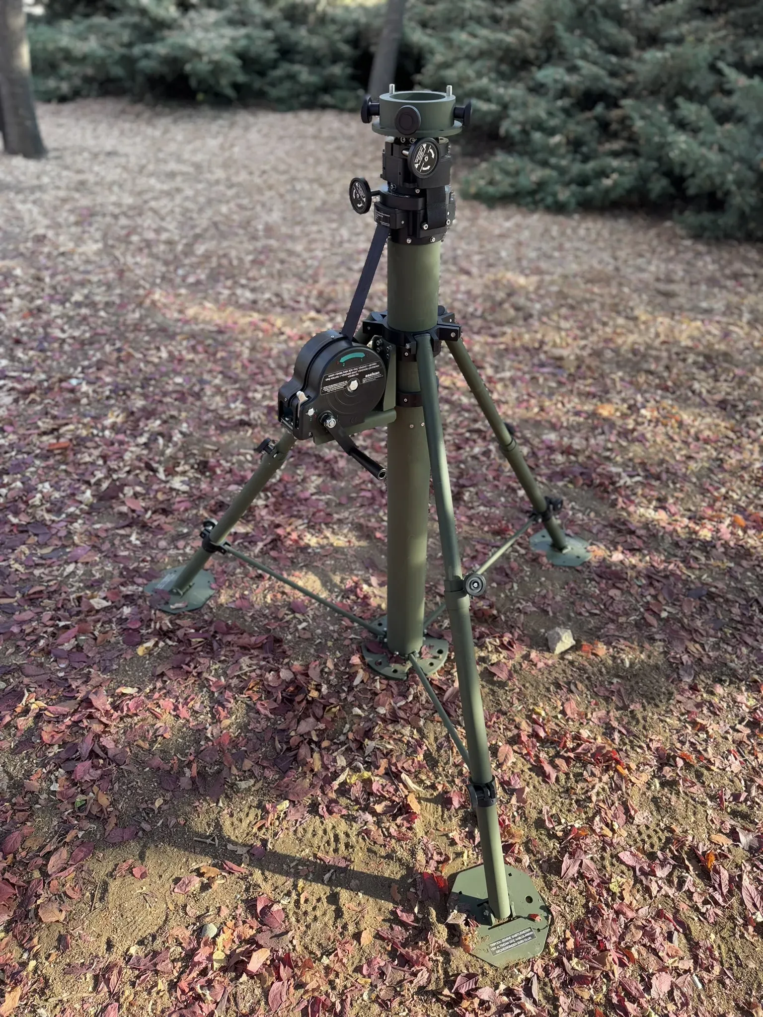 Crane-arm mast stowed on its splayed tripod on leaf-covered ground in front of evergreens.