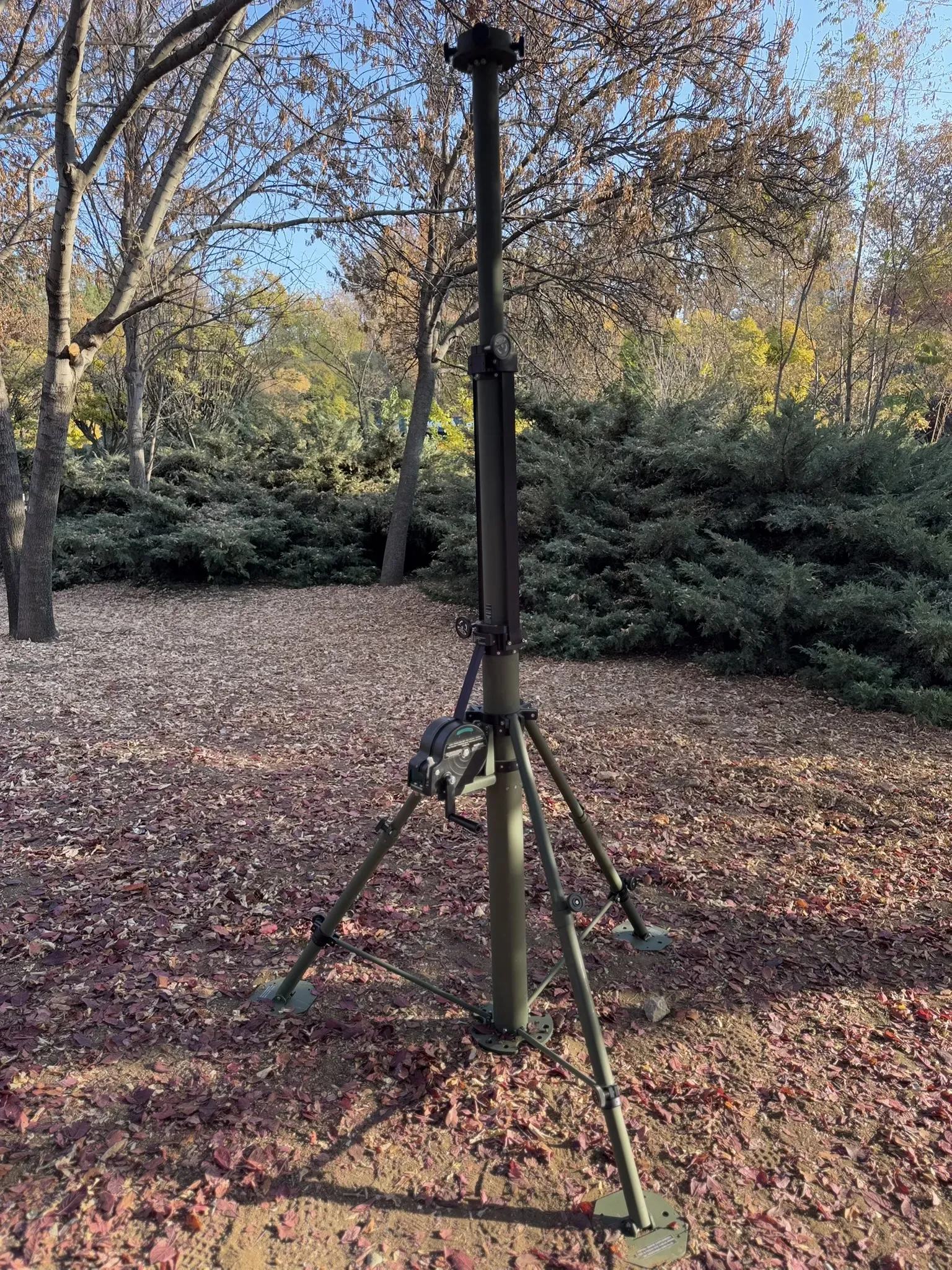 Partly raised olive mast on tripod standing in open woodland with bare autumn trees behind.
