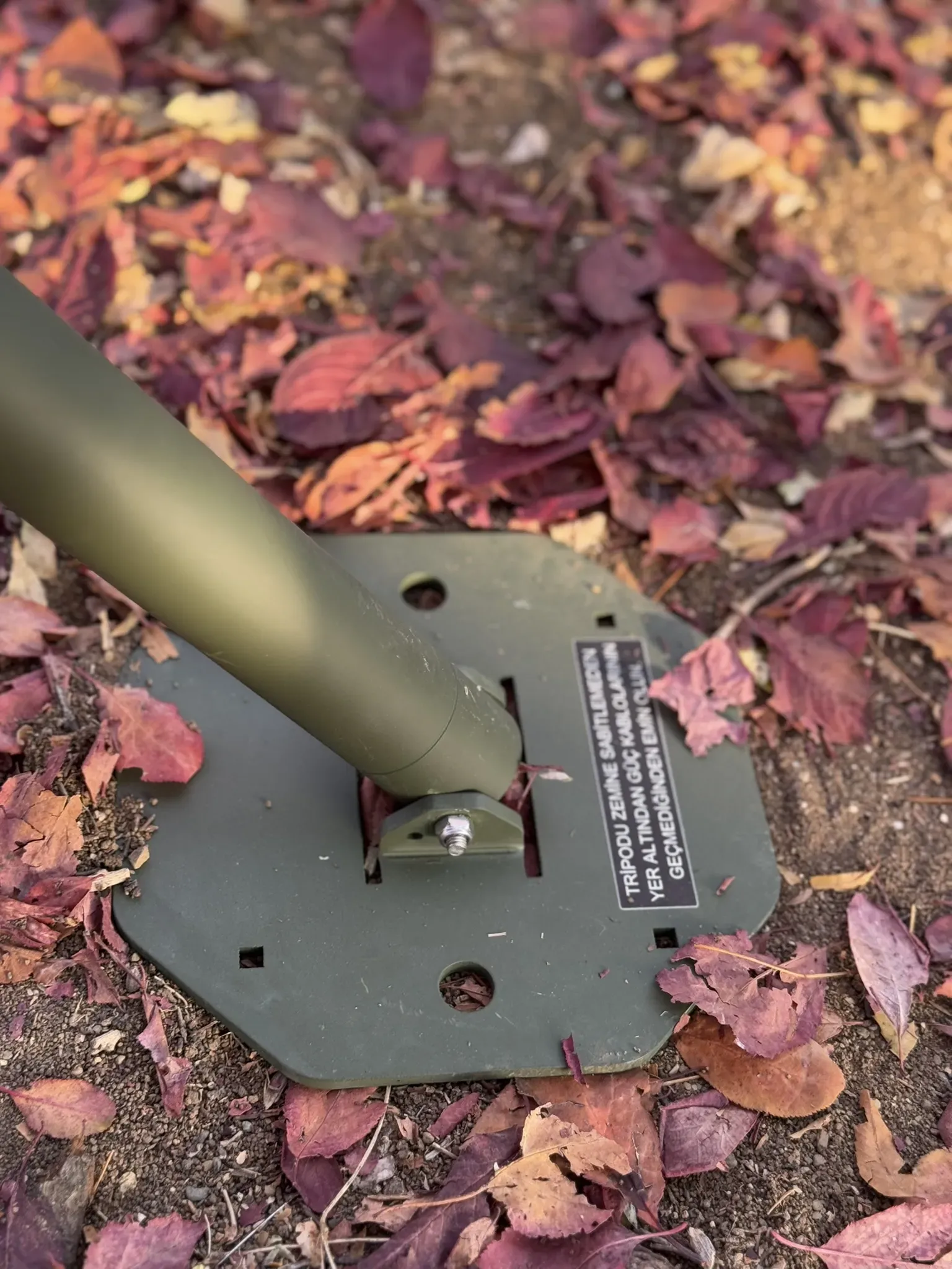 Tripod leg bolted into its olive octagonal footplate resting on red fallen leaves.