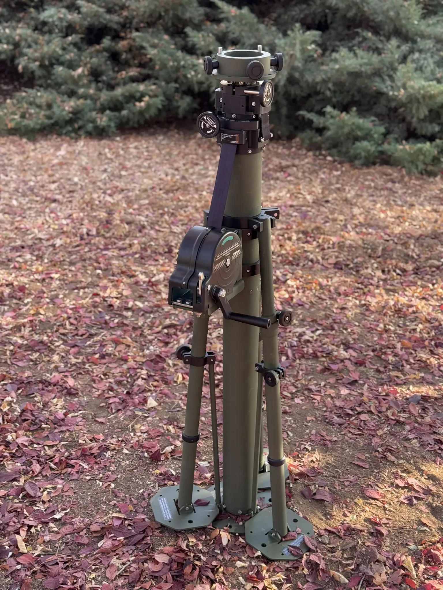Stowed crane-arm mast on tripod with winch on the pole, evergreen hedge behind.