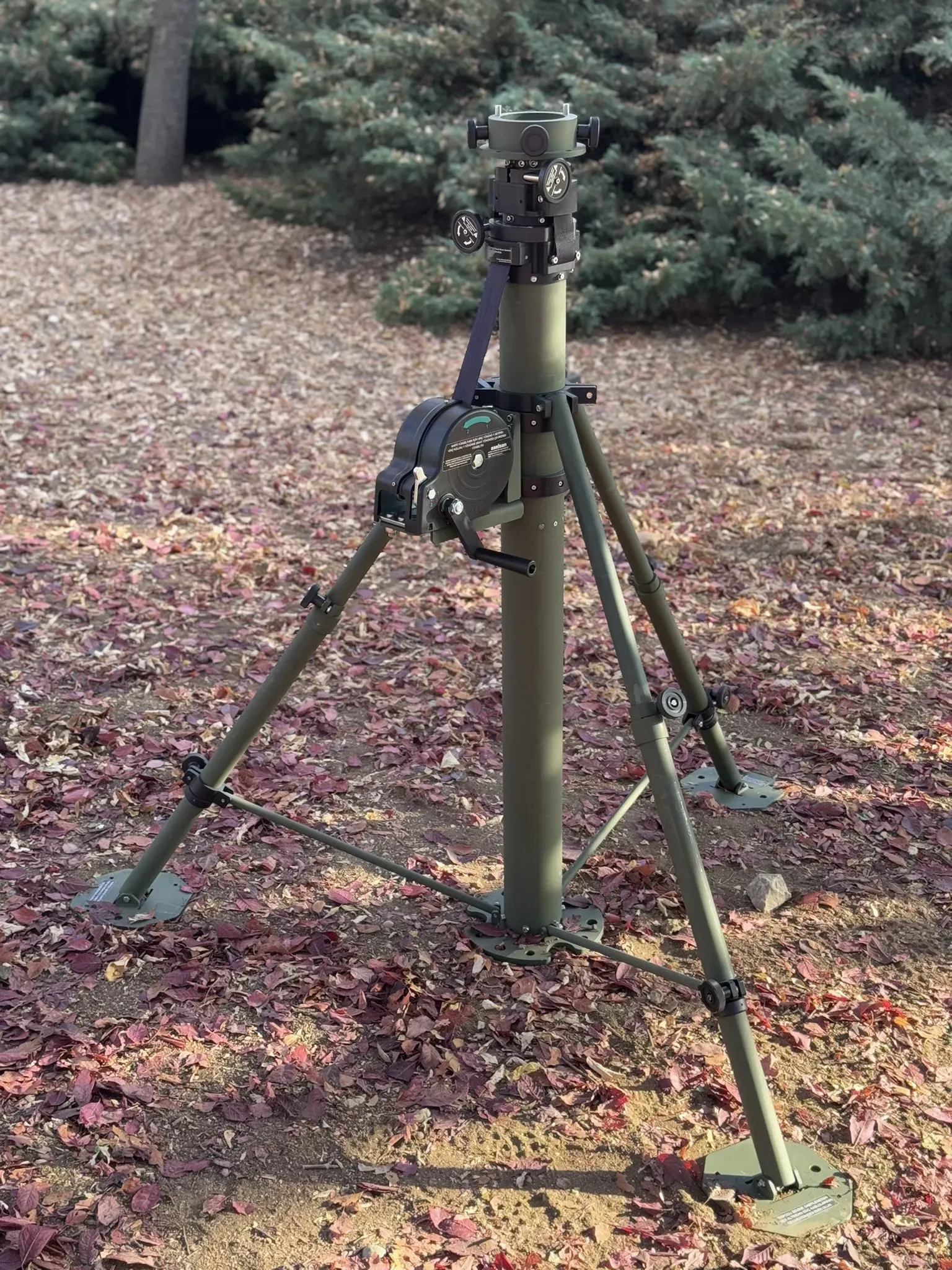 Stowed mast with widely splayed tripod legs and footplates on leaf-covered woodland ground.