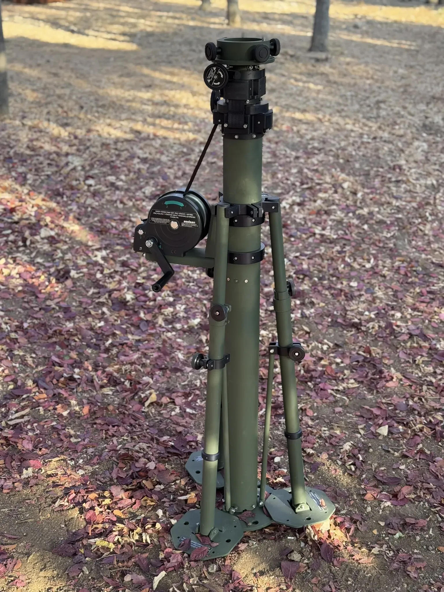 Stowed mast on tripod with side winch, standing on a sunlit leaf-strewn woodland path.
