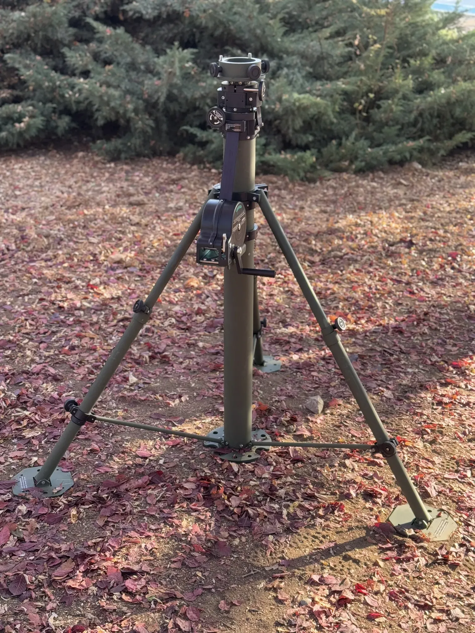 Crane-arm mast stowed on its tripod on leaf-covered ground with dense evergreens behind.