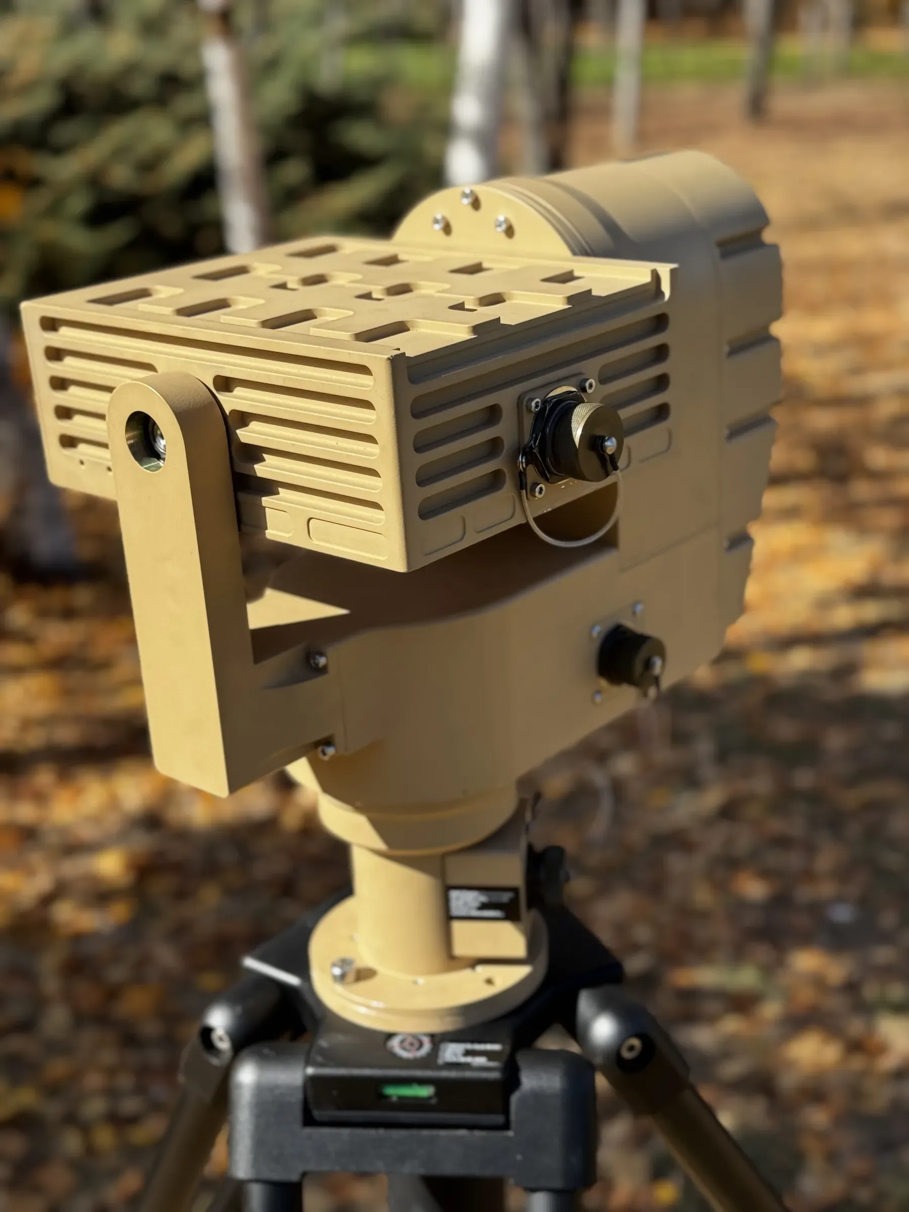 Close low-angle view of the tan PED12 positioner head and finned payload housing, tripod head visible below.