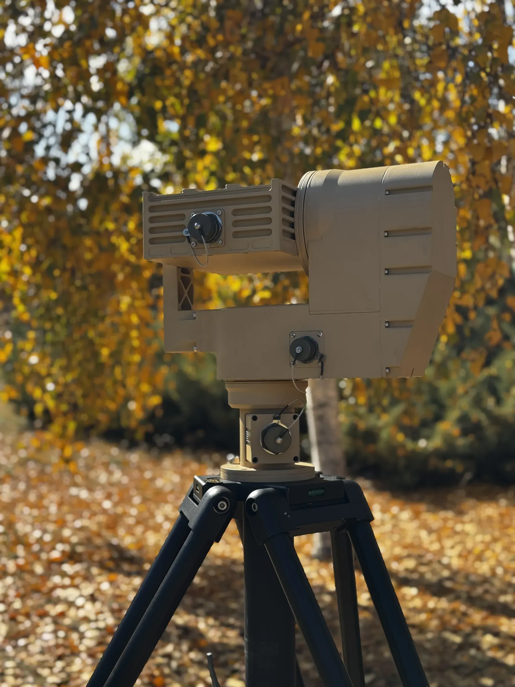 Tan PED12 positioner on black tripod seen from the rear left, yellow autumn foliage and sky behind.