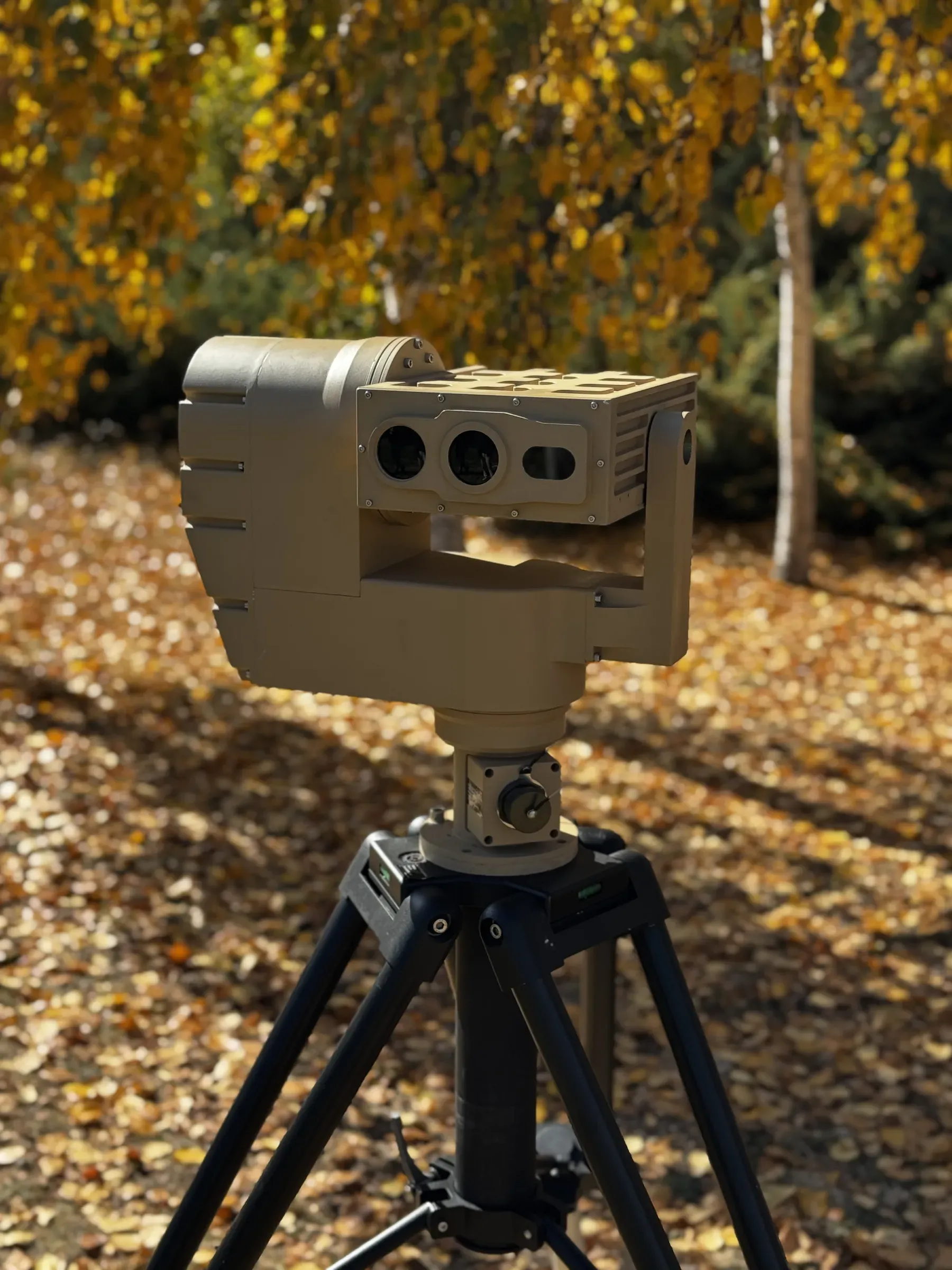 Tan PED12 positioner with three-window optics head on a black tripod, framed against golden autumn birch trees.