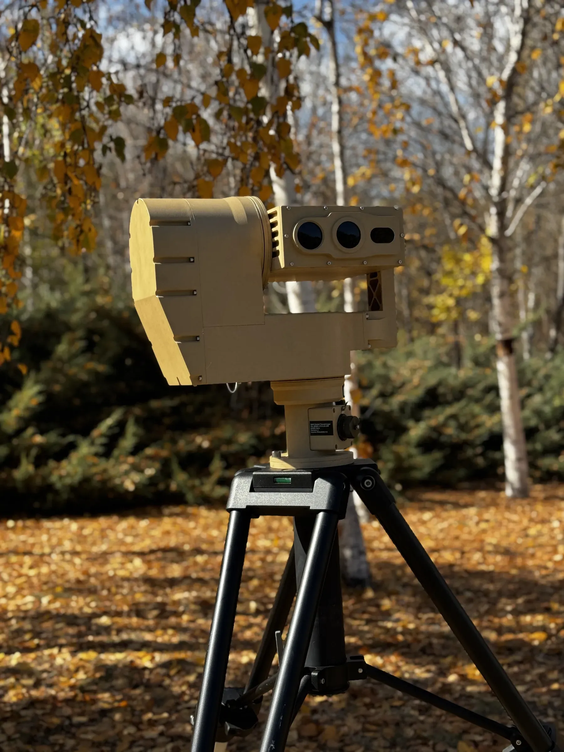 Low-angle shot of the tan PED12 positioner and its optics head on a black tripod amid autumn undergrowth.