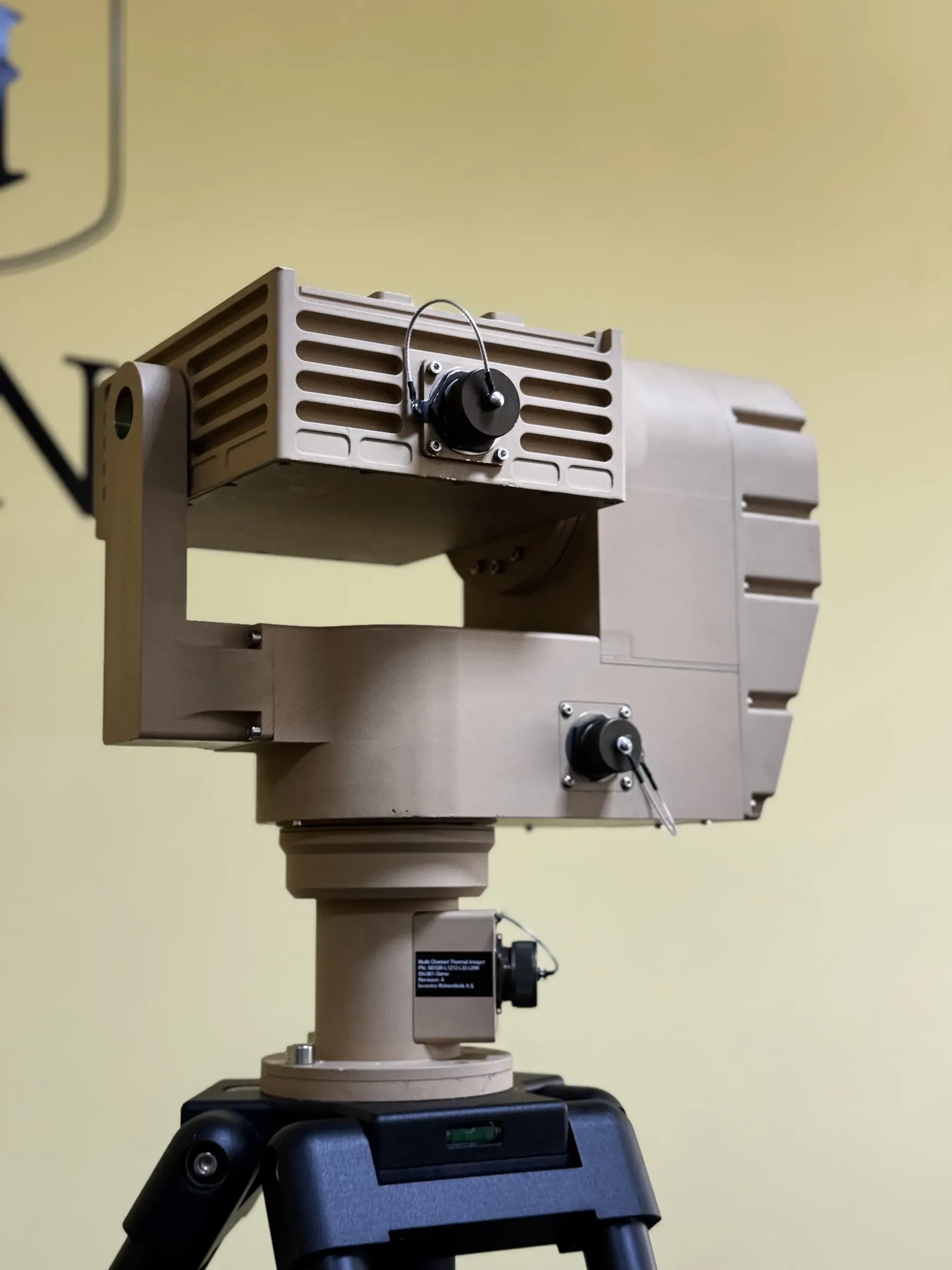 Tan PED12 pan-tilt positioner carrying a SEB20 thermal module on a tripod, photographed from a low three-quarter angle indoors.