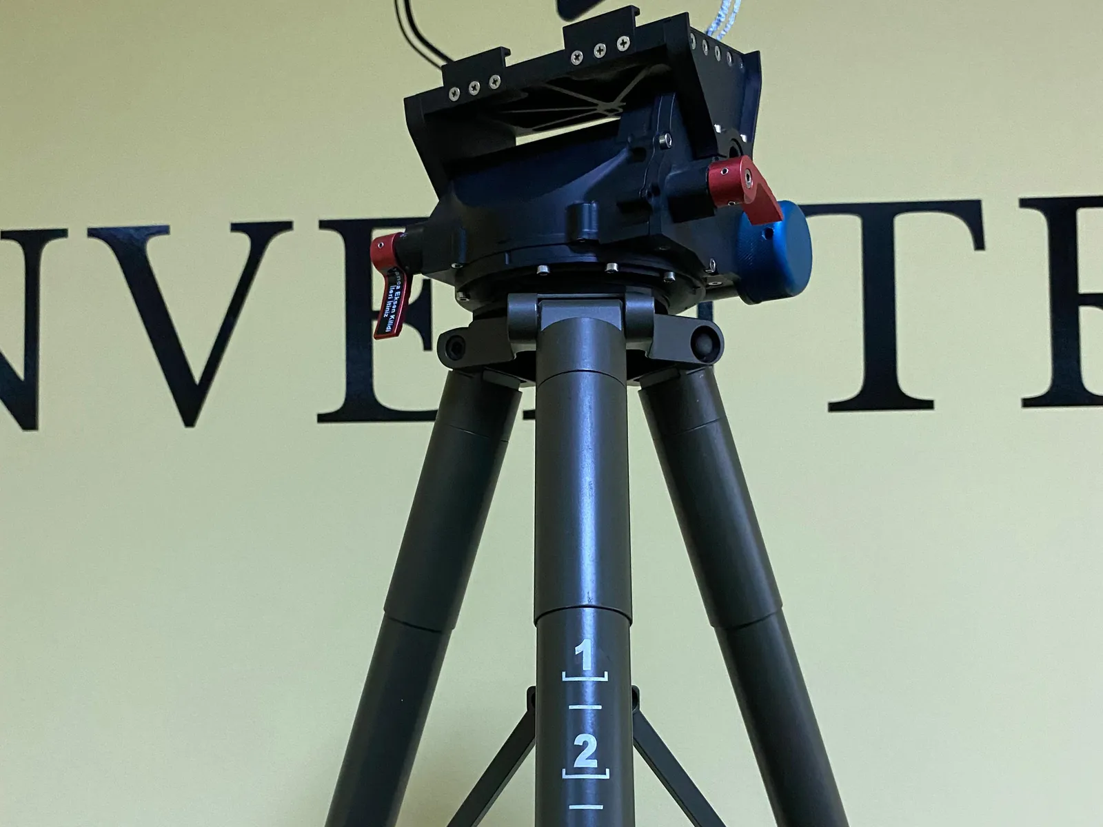 Lightweight manual pan-tilt head on a tripod column at left-front three-quarter: red pan lock lever, blue tilt lock disc and payload plate