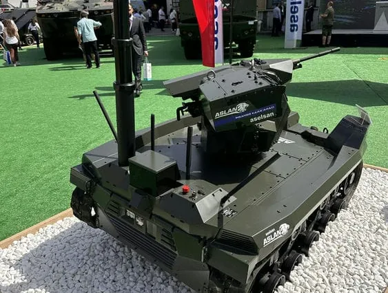 MES01 mast with SEG10 gimbal raised on an ASELSAN tracked UGV at the IDEF 2025 outdoor stand, visitors nearby.