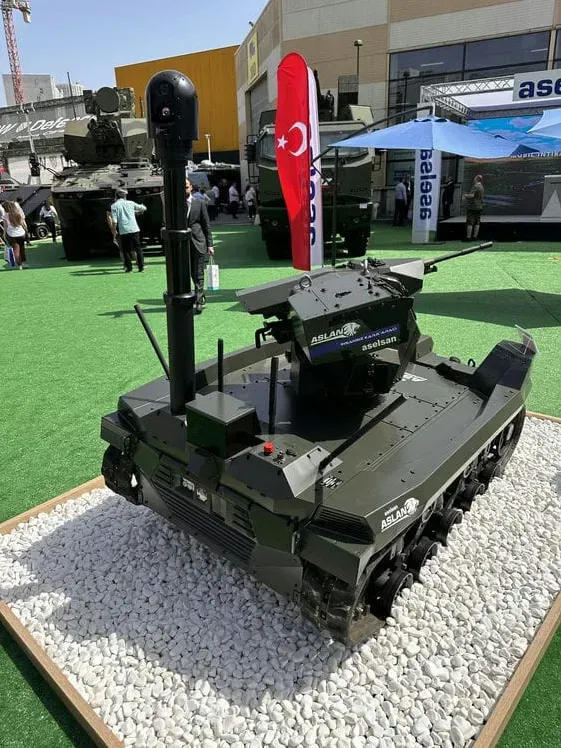 Dark green tracked ASELSAN UGV with the MES01 mast and SEG10 head stowed, displayed outdoors on a gravel bed.