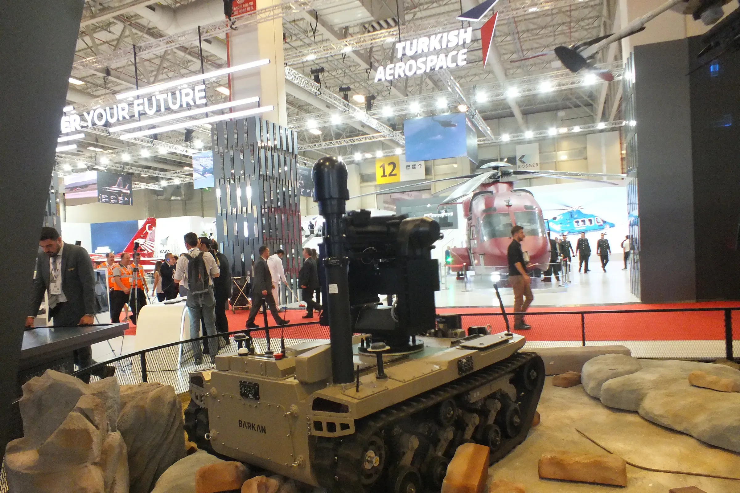 Tan tracked vehicle with the MES01 mast raised, shown on a rock diorama inside the IDEF hall with a helicopter behind.