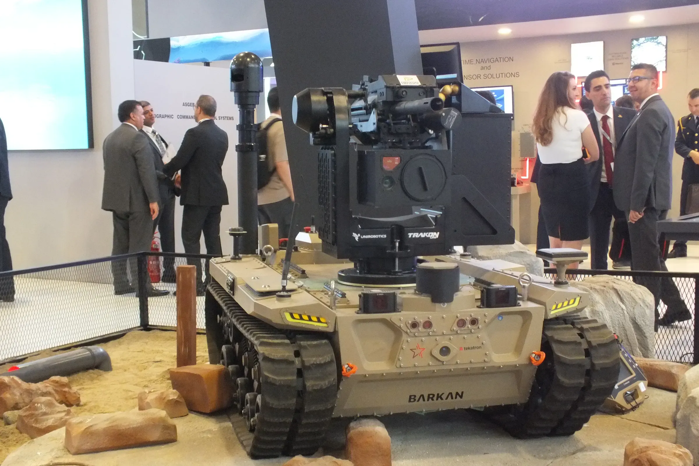 Sand-coloured BARKAN tracked UGV on a rock diorama with the MES01 mast and payload raised, visitors around it.