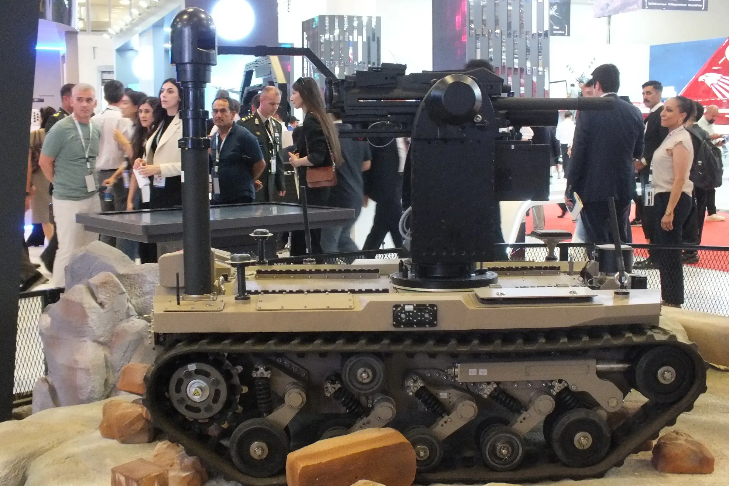 Rear view of tracked unmanned ground vehicle with mast raised beside its weapon station, visitors crowding the aisle behind.
