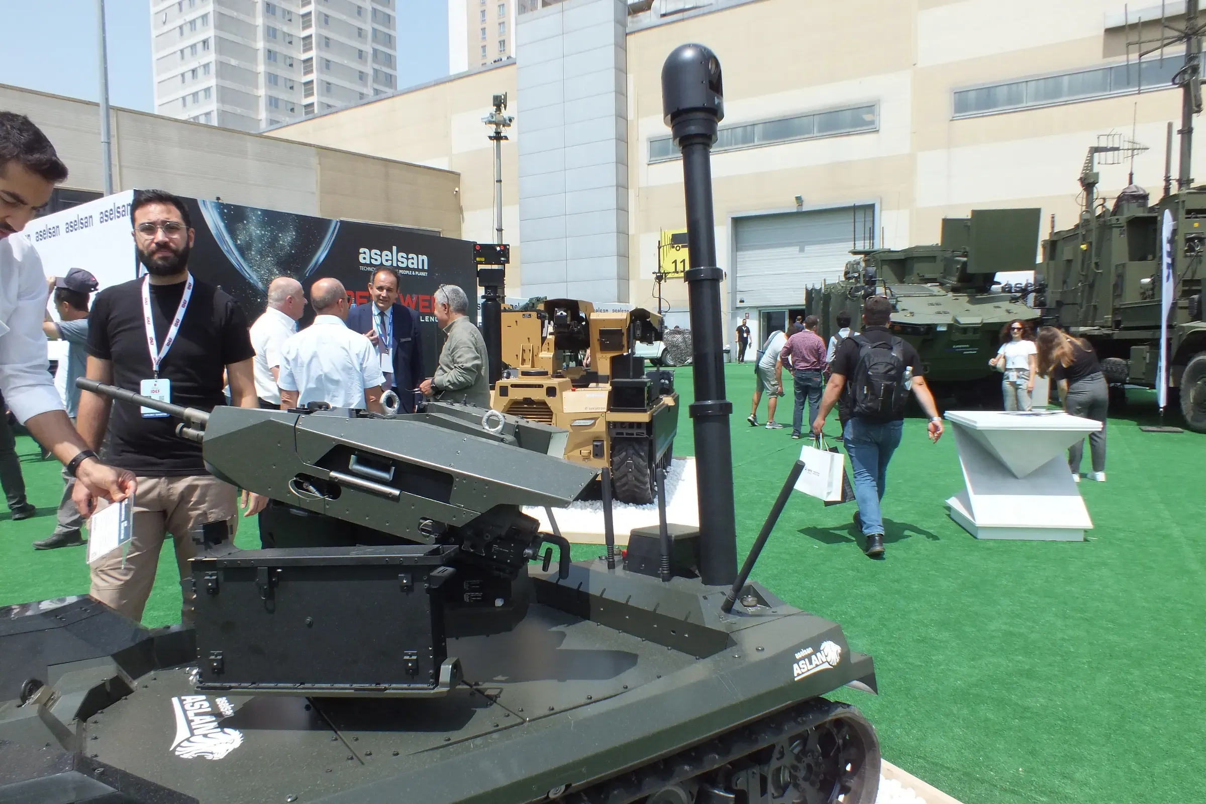 Green tracked vehicle with fully extended mast at an outdoor stand, exhibitor standing left, armoured vehicles and visitors behind.
