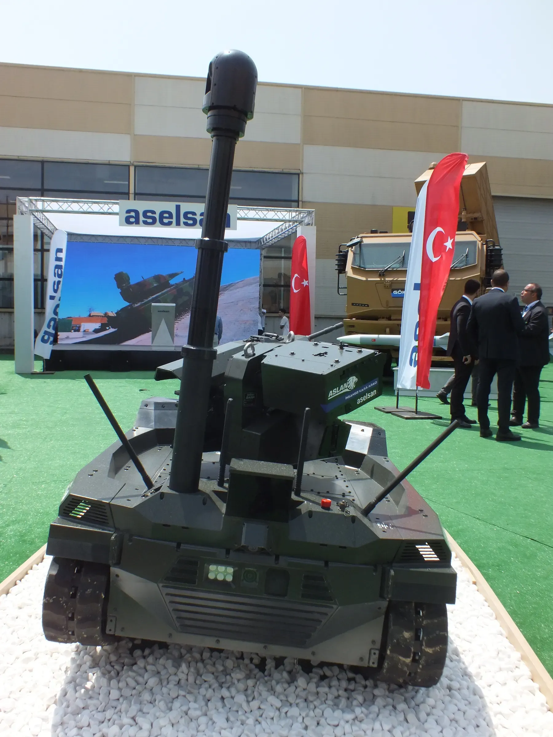 Front view of green tracked vehicle on a gravel plinth, mast extended vertically, ASELSAN banner and Turkish flag behind.