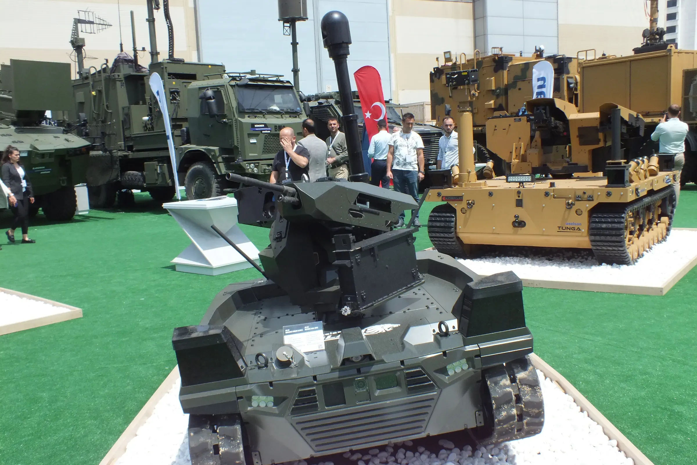 Three-quarter front view of green tracked vehicle on gravel with mast raised, tan tracked vehicle and trucks alongside.