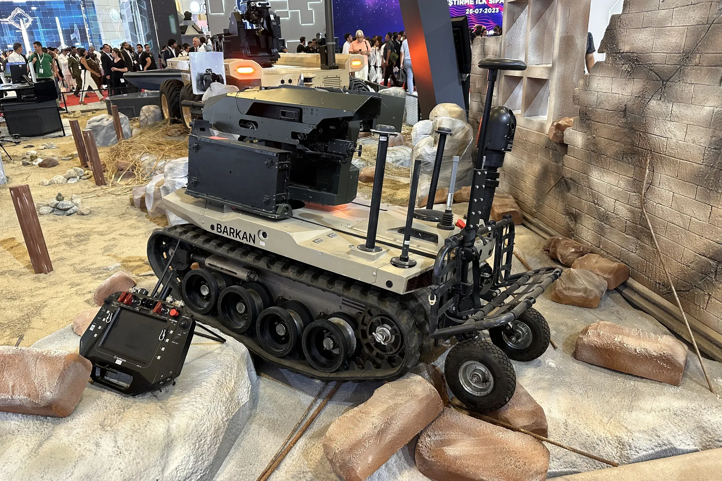 Tan BARKAN tracked vehicle on artificial rocks inside the hall, mast extended at rear, control unit lying in front.