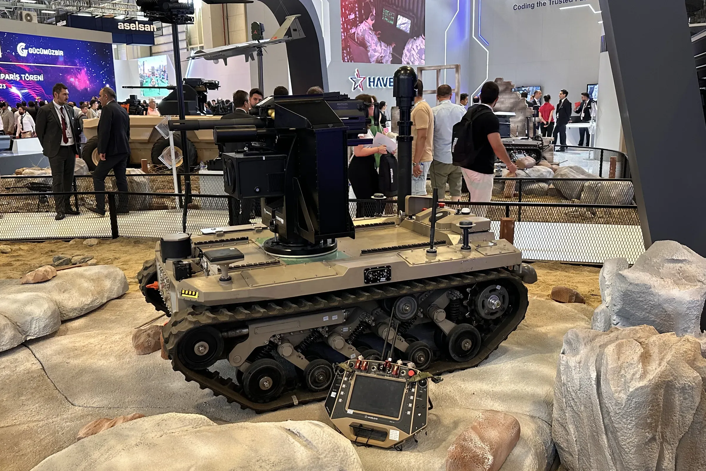 Side view of tan tracked vehicle on rocks with mast and turret raised, HAVELSAN and ASELSAN stands behind.