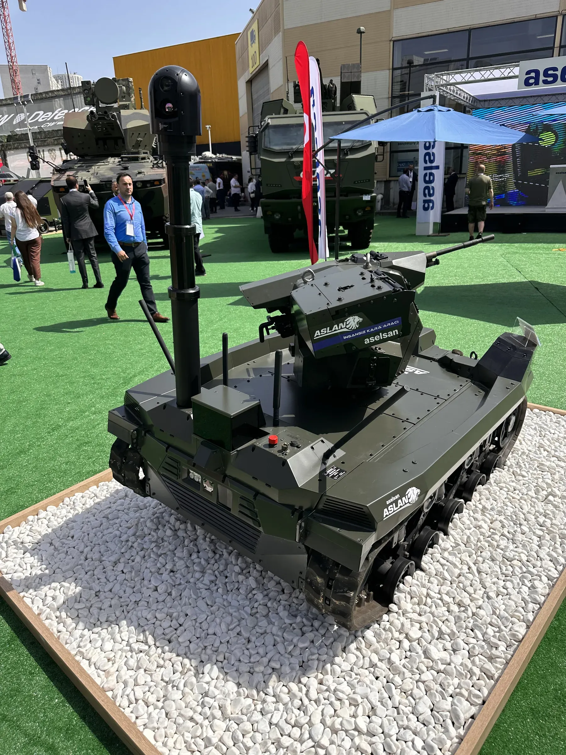 Green tracked vehicle on a white gravel plinth with mast extended, ASELSAN banners and visitors at the outdoor stand.