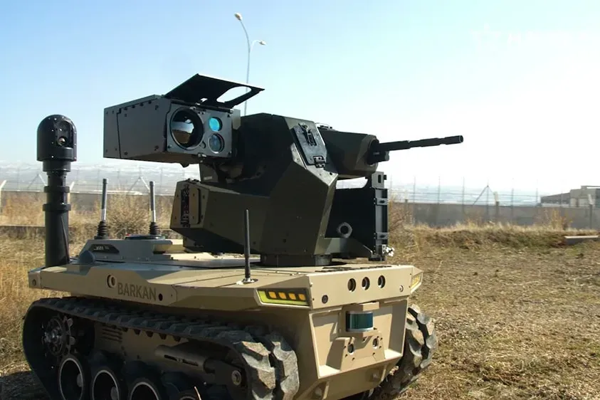 Tan tracked vehicle with extended mast and turret standing on dry ground, perimeter fence in the distance.