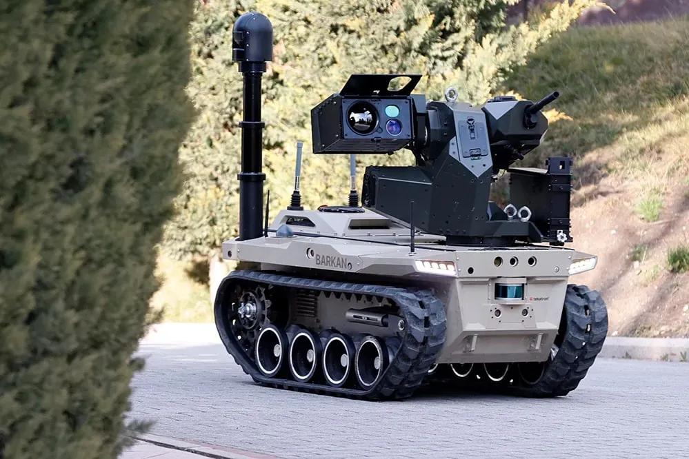 Tan tracked vehicle with raised mast and sensor turret on paving beside a steep vegetated rock face.