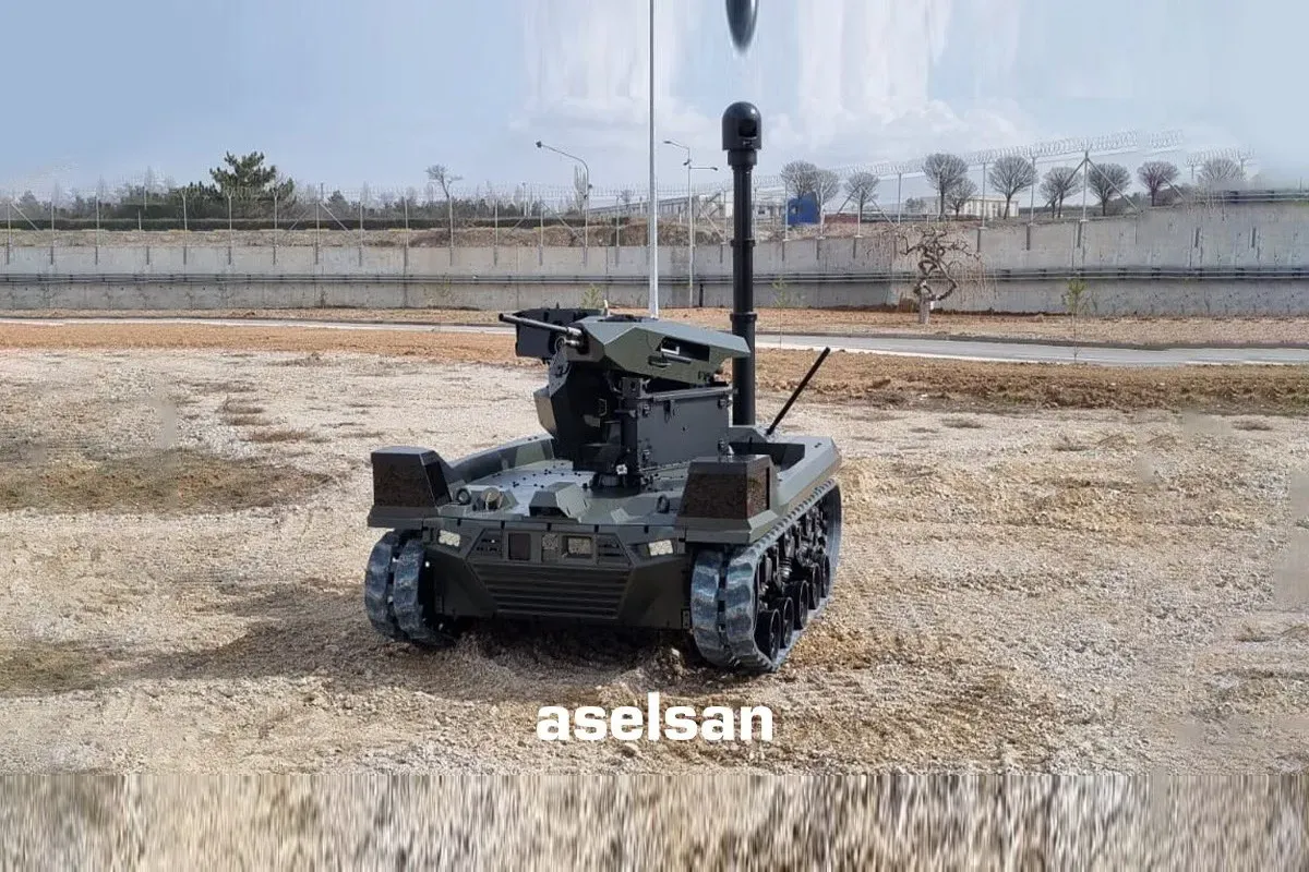 Green tracked vehicle with raised mast and turret on bare earth, perimeter wall behind and aselsan watermark.