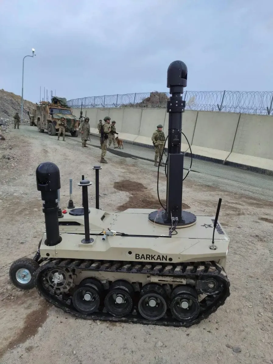 Tan BARKAN UGV fitted with two raised mast gimbals, soldiers and vehicles behind a wall.