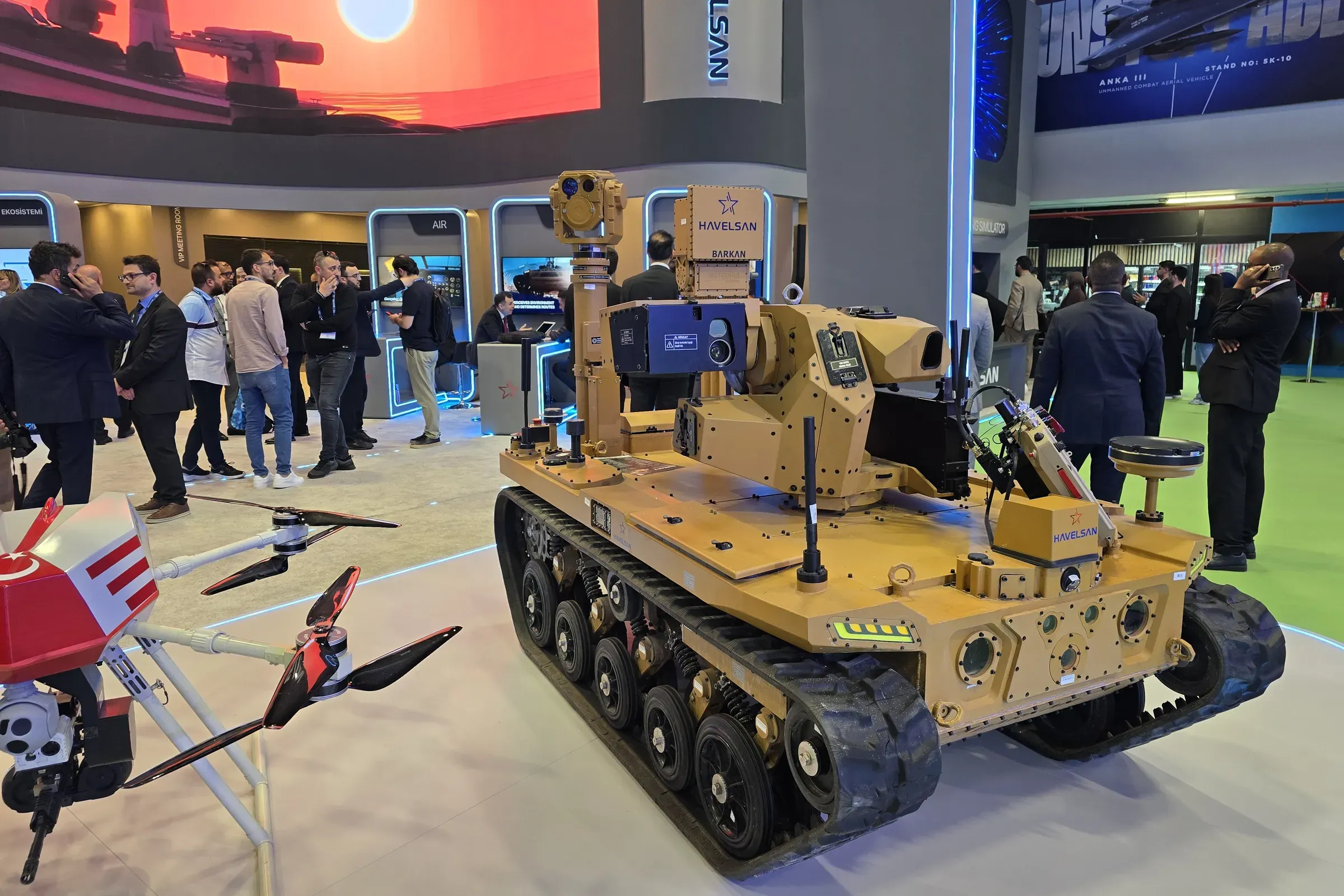 Tan tracked BARKAN UGV carrying the MEM10 mast and SEG14 gimbal on the HAVELSAN stand, visitors around it.
