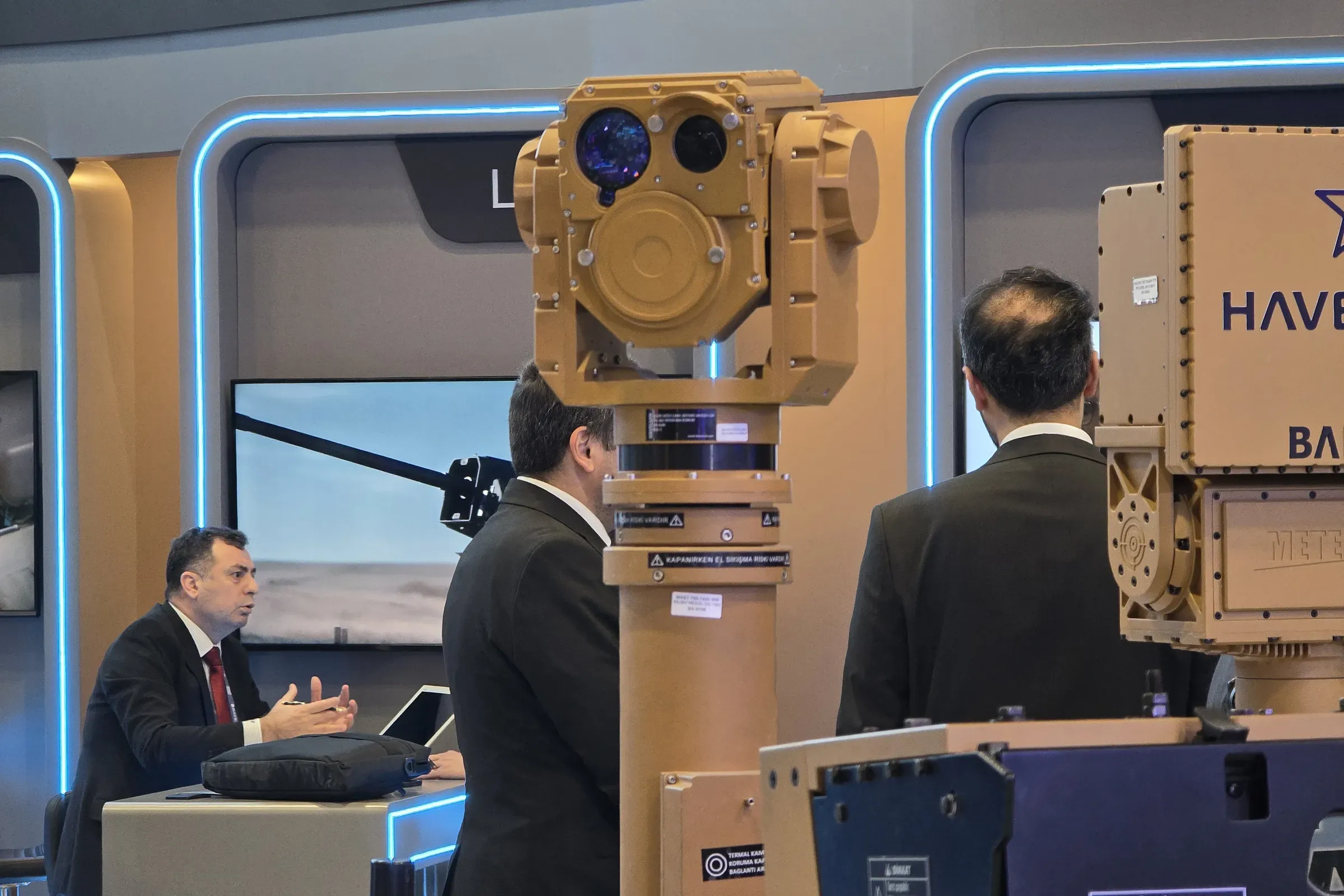 Close view of the SEG14 gimbal head on the extended tan mast, visitors talking at the HAVELSAN stand below.