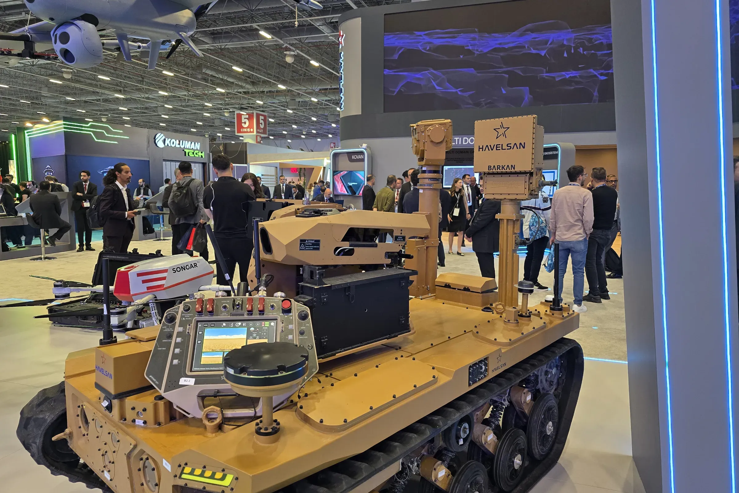 Tan tracked BARKAN UGV with stowed MEM10 mast, viewed from the front on the stand with a UAV suspended above.