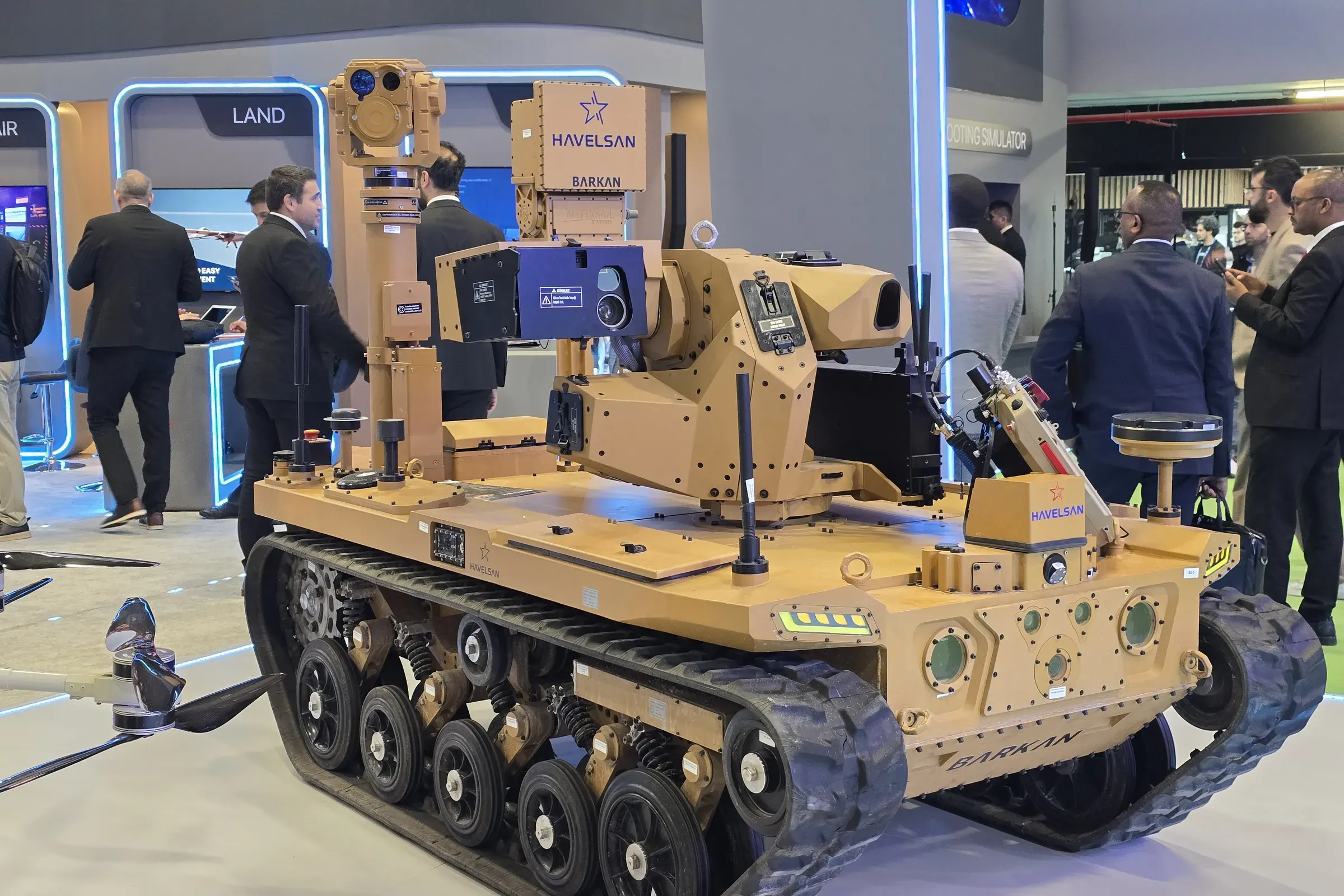 Side view of the tan BARKAN tracked UGV with the MEM10 mast and gimbal erected, crowd on the stand behind.