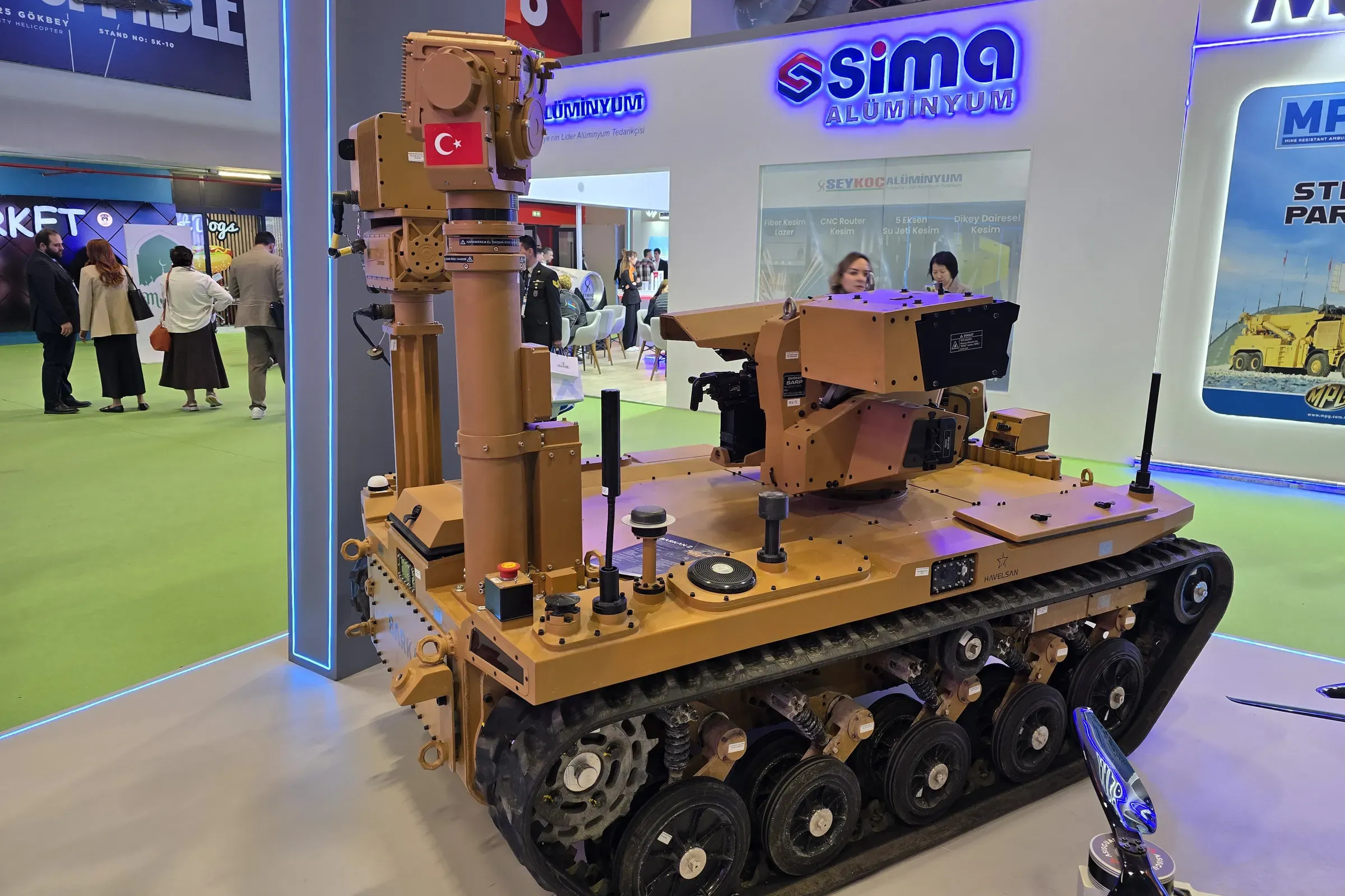 Tan tracked UGV with the MEM10 mast partly extended, seen head-on across the green exhibition carpet.