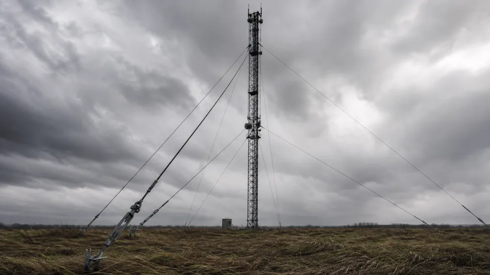 A guyed lattice mast against a heavy storm sky, its diagonal guy cables held by tensioners and ground anchors above wind-flattened grass.